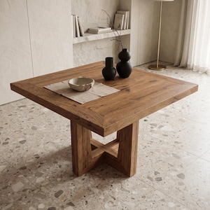 Op de afbeelding: Een vierkante houten eettafel met een dik blad en een stevig kruisvormig onderstel. Op de tafel liggen een lichtgekleurde vierkante stoffen placemat, een ondiepe keramische kom en twee matzwarte vazen van verschillende hoogtes.
