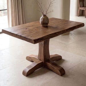 May include: A rectangular wooden dining table with a thick, natural live-edge top and a sturdy central pedestal base featuring four curved feet. A small, round, textured ceramic vase containing thin, bare branches sits on the table surface.