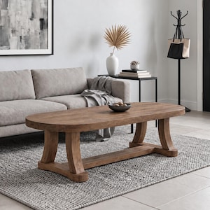 May include: A light brown wooden coffee table with an oval top and curved legs, positioned on a gray textured rug in front of a light gray sofa. The table features a horizontal support beam connecting the four legs, providing a stable base.