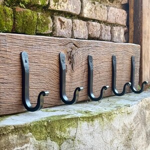 May include: A wooden coat rack with five black metal hooks. The wood has a natural, weathered appearance. The hooks are evenly spaced and have a curved design. The rack is mounted on a stone wall.