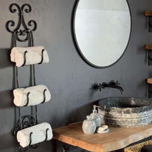 Può includere: Portasciugamani in metallo nero a tre livelli, con asciugamani bianchi arrotolati. Il portasciugamani ha un design ornato e arricciato nella parte superiore. Uno specchio rotondo e una parete grigio scuro sono sullo sfondo. È visibile anche un piano di lavoro in legno con un lavabo in pietra.