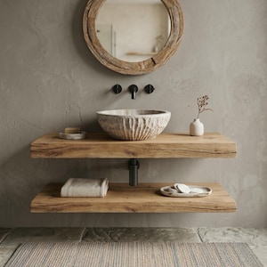 Può includere: Un mobile da bagno rustico con un lavabo in pietra scolpita, mensole in legno e uno specchio rotondo in legno. Le mensole contengono una saponetta, un asciugamano arrotolato e dischetti di cotone. Il rubinetto è nero e il muro è grigio neutro.