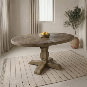 Op de afbeelding: Een grote, ronde houten eettafel met een voetstuk. De tafel is gemaakt van verweerd hout en heeft een natuurlijke afwerking. Een kom met citroenen staat op het tafelblad. De tafel staat op een gestreept tapijt.