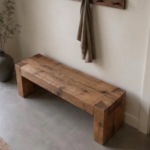 May include: A rectangular wooden bench constructed from thick, reclaimed timber planks. The bench features a natural brown wood grain finish with visible knots and joinery details at the corners where the legs meet the seat, creating a sturdy, block-like form.