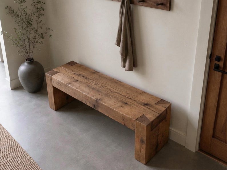 May include: A rectangular wooden bench constructed from thick, reclaimed timber planks. The bench features a natural brown wood grain finish with visible knots and joinery details at the corners where the legs meet the seat, creating a sturdy, block-like form.
