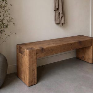 May include: A rectangular wooden bench constructed from thick, solid timber planks with a natural brown finish. The bench features a simple, block-style design with four sturdy legs integrated into the frame, showcasing a visible wood grain texture.