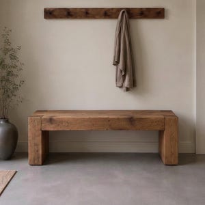 May include: A rectangular wooden bench with a natural grain finish, featuring thick block legs. Above the bench, a wooden wall-mounted coat rack holds a single beige fabric garment. A dark grey ceramic vase with thin branches sits to the left.