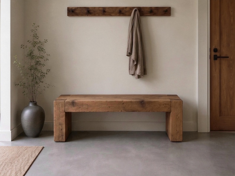 May include: A rectangular wooden bench with a natural grain finish, featuring thick block legs. Above the bench, a wooden wall-mounted coat rack holds a single beige fabric garment. A dark grey ceramic vase with thin branches sits to the left.