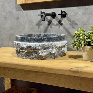 May include: A round, dark grey stone sink with a rough, textured exterior sits on a wooden countertop. A black faucet is mounted on the wall behind the sink.
