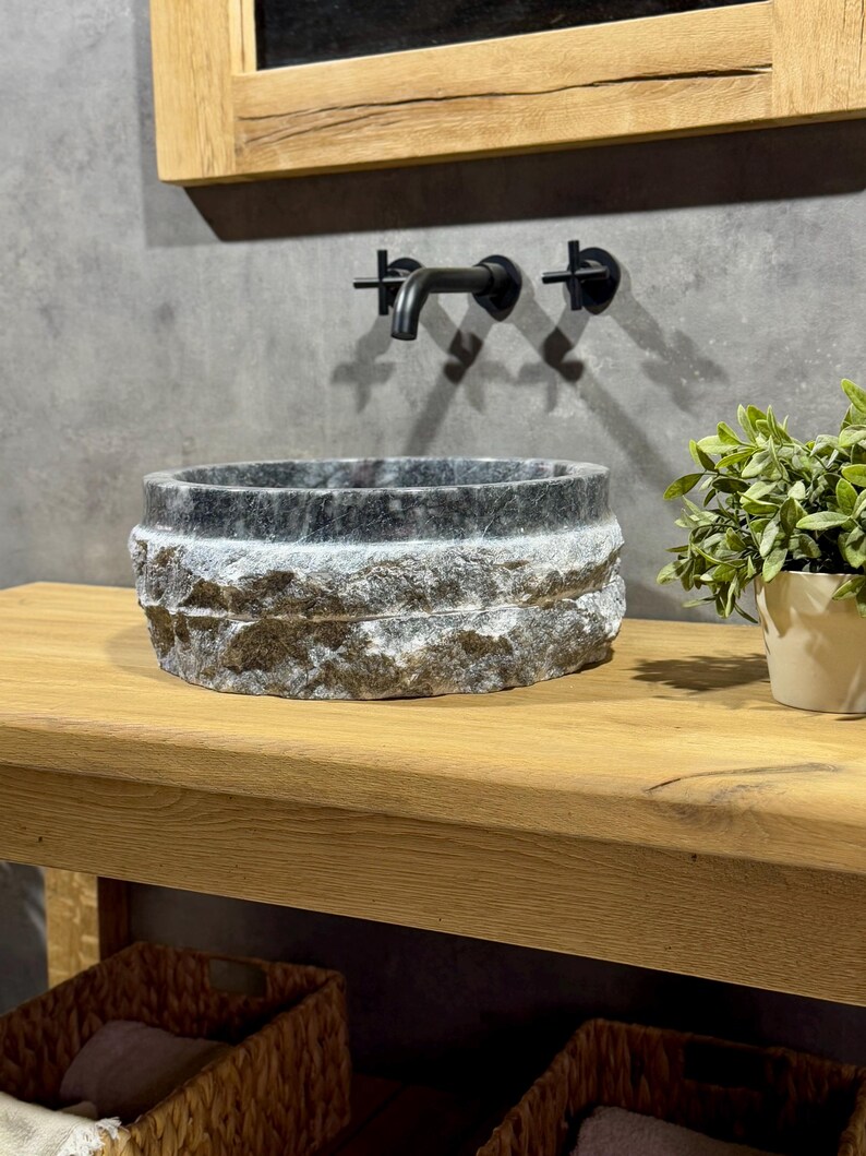 May include: A round, dark grey stone sink with a rough, textured exterior sits on a wooden countertop. A black faucet is mounted on the wall behind the sink.