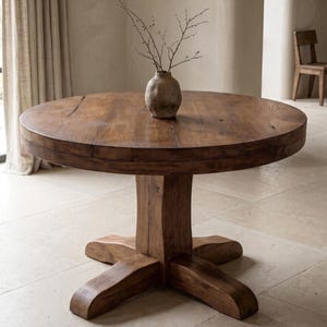 May include: A round, dark brown wooden dining table with a thick, circular top and a sturdy pedestal base featuring four wide, extending feet. A small, textured, light brown ceramic vase containing thin, bare branches sits in the center.