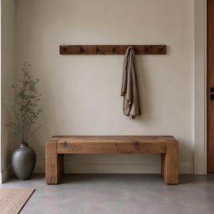 May include: A rectangular wooden bench with a thick, solid construction and a natural brown wood grain finish. Above the bench, a matching horizontal wooden wall-mounted coat rack holds a single beige fabric garment hanging from a hook.