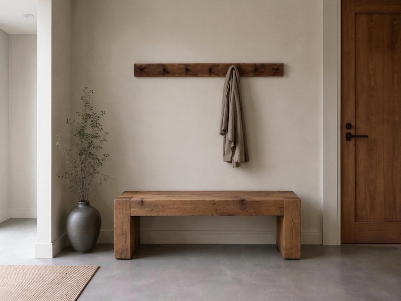 May include: A rectangular wooden bench with a thick, solid construction and a natural brown wood grain finish. Above the bench, a matching horizontal wooden wall-mounted coat rack holds a single beige fabric garment hanging from a hook.