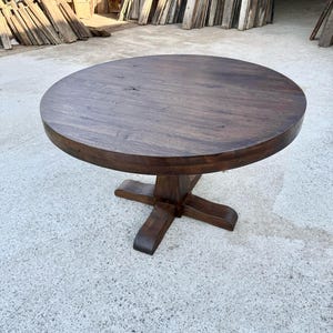 May include: A round, dark brown wooden dining table with a smooth, circular top and a central pedestal base featuring four sturdy, outward-extending feet. The wood grain is visible across the surface, showcasing a deep, rich, stained finish.
