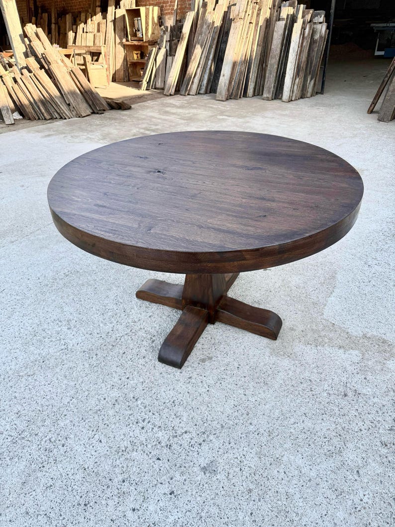 May include: A round, dark brown wooden dining table with a smooth, circular top and a central pedestal base featuring four sturdy, outward-extending feet. The wood grain is visible across the surface, showcasing a deep, rich, stained finish.