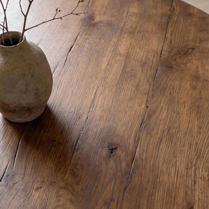 May include: A round wooden table with a dark brown finish, visible wood grain, and natural knots. A small, textured, light-colored ceramic vase containing a thin, bare branch with tiny buds sits on the surface of the table.