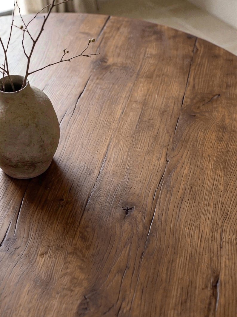 May include: A round wooden table with a dark brown finish, visible wood grain, and natural knots. A small, textured, light-colored ceramic vase containing a thin, bare branch with tiny buds sits on the surface of the table.