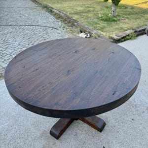 May include: A round dining table with a dark brown wood grain finish. The tabletop features visible natural wood knots and texture. It is supported by a central pedestal base that branches out into a sturdy, four-legged cross design at the bottom.