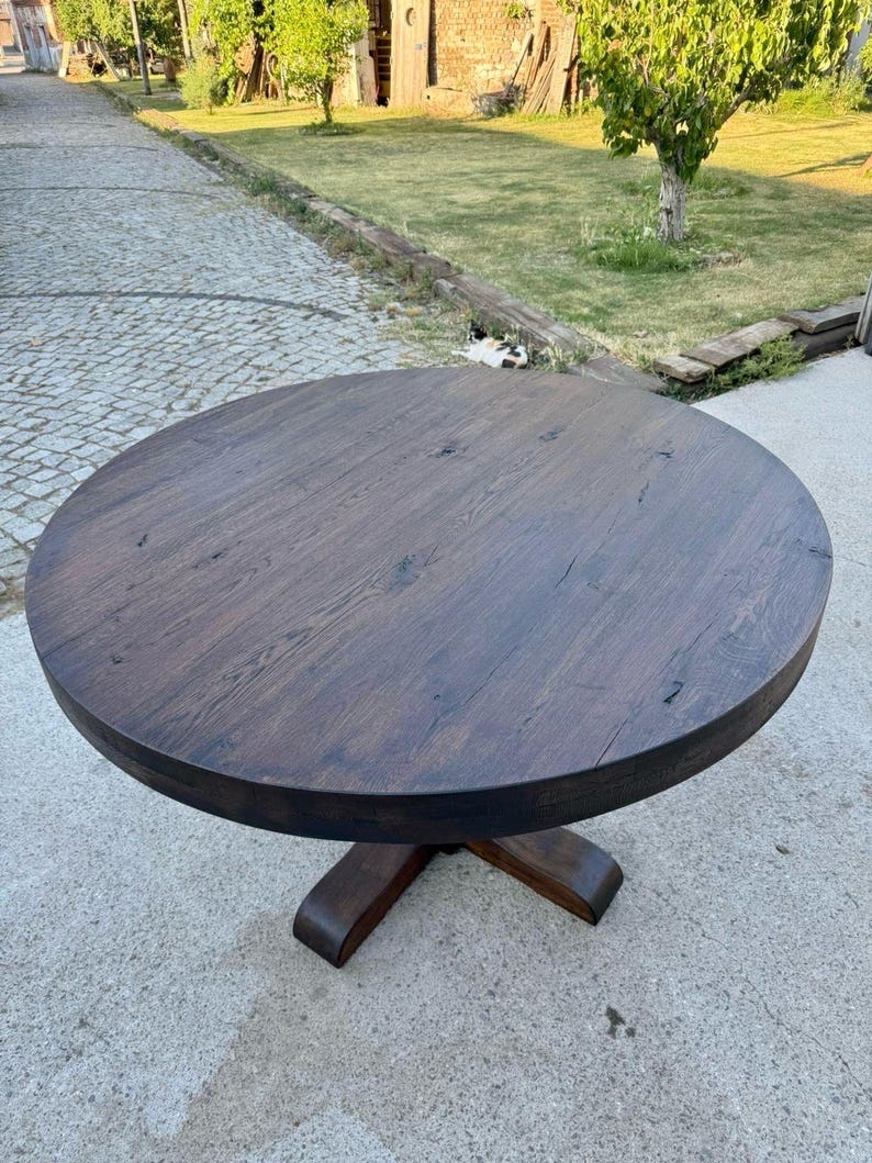 May include: A round dining table with a dark brown wood grain finish. The tabletop features visible natural wood knots and texture. It is supported by a central pedestal base that branches out into a sturdy, four-legged cross design at the bottom.