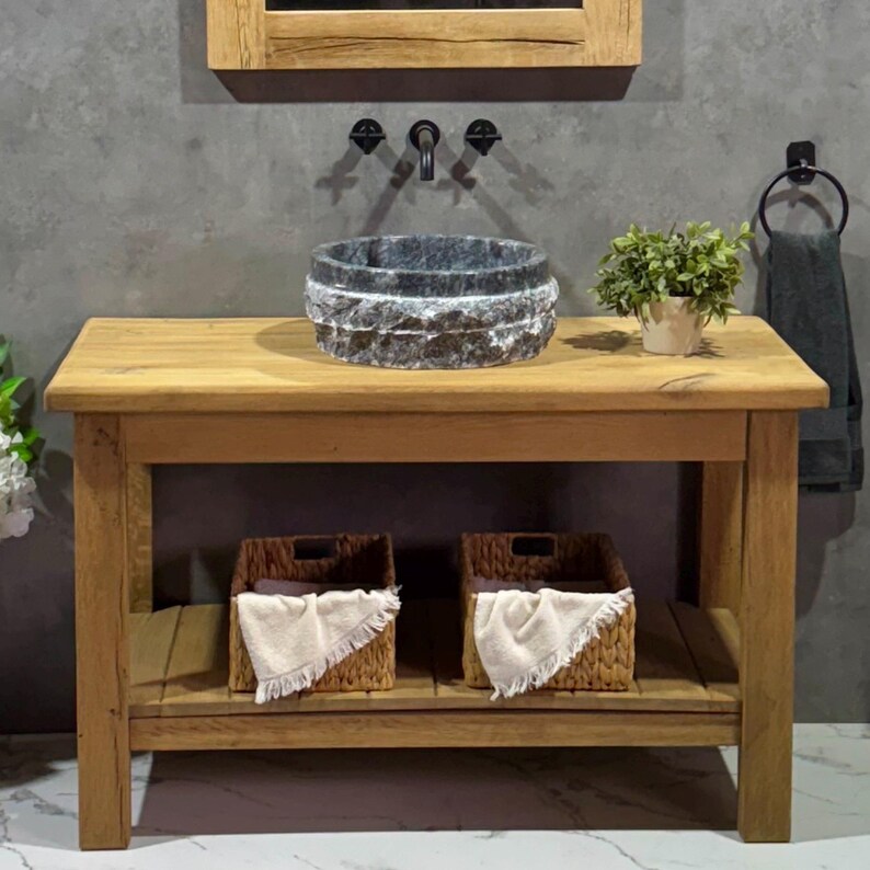 May include: A rustic wooden bathroom vanity with a black stone vessel sink, two woven baskets, and a small potted plant. The vanity has a shelf for storage.