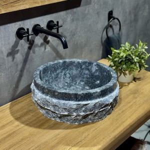 May include: A dark grey stone vessel sink with a rough, textured exterior. The sink is mounted on a wooden countertop.