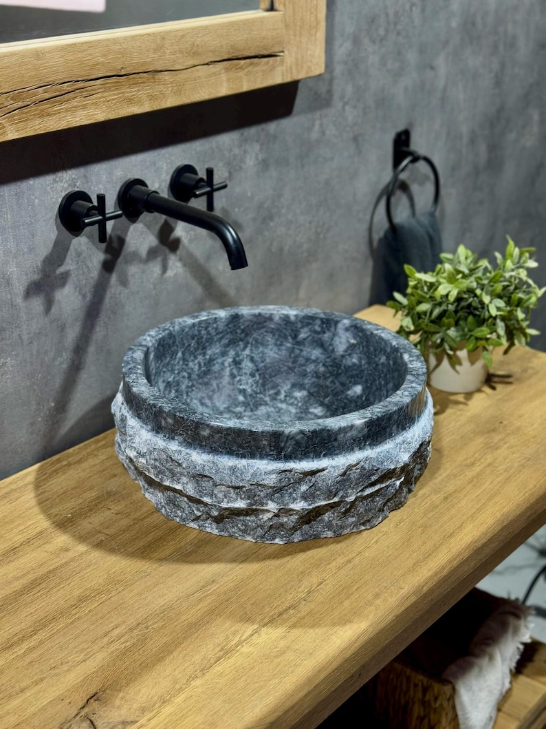 May include: A dark grey stone vessel sink with a rough, textured exterior. The sink is mounted on a wooden countertop.