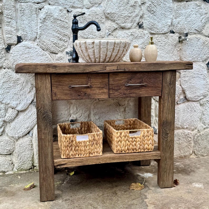 May include: A rustic wooden bathroom vanity with two drawers and a lower shelf. The vanity features a white ceramic vessel sink with a black faucet and two woven baskets on the shelf.