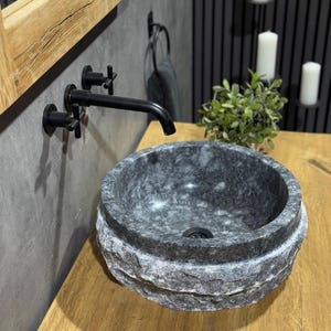 May include: A round, grey stone sink with a rough, textured exterior. The sink is mounted on a wooden countertop and has a black faucet with a cross-shaped handle.
