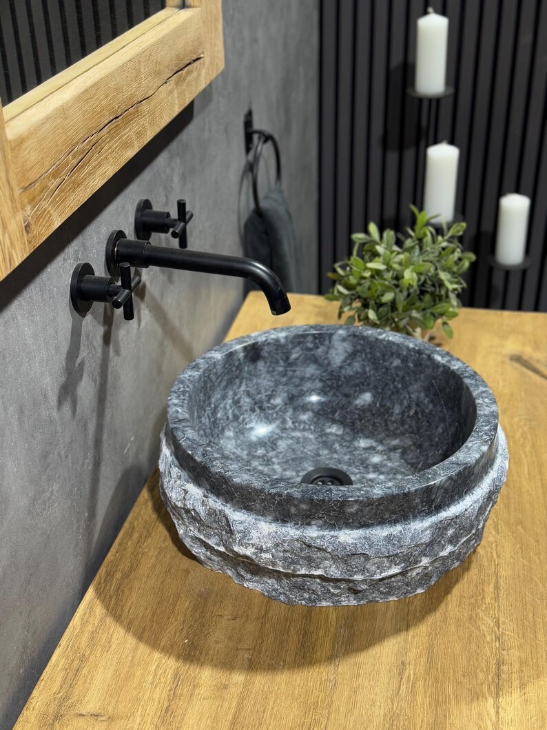 May include: A round, grey stone sink with a rough, textured exterior. The sink is mounted on a wooden countertop and has a black faucet with a cross-shaped handle.
