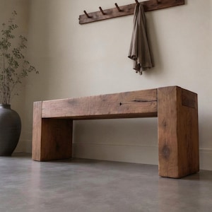 May include: A rectangular bench constructed from thick, reclaimed brown timber with a visible wood grain and natural knots. The bench features a sturdy, block-style design with two wide vertical supports and a thick horizontal seat surface.