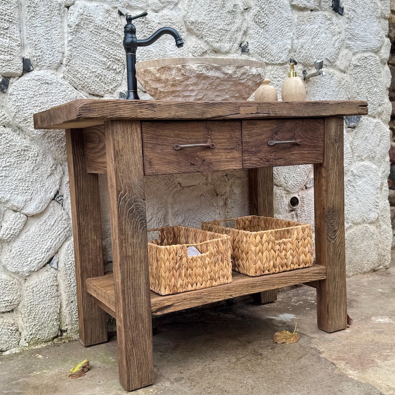 May include: A rustic wooden bathroom vanity with two drawers and a lower shelf. The vanity is made of reclaimed wood and has a natural finish. It features a stone sink and a black faucet. Two woven baskets are on the lower shelf.