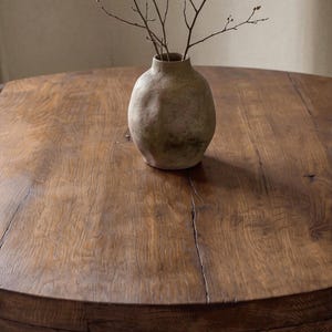 May include: A round, dark brown wooden table with a visible grain and natural cracks, featuring a textured, beige, bulbous ceramic vase at the center. The vase contains a few thin, bare, brown branches extending upward from its narrow opening.