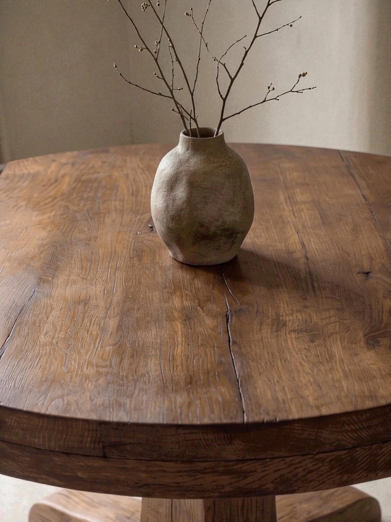 May include: A round, dark brown wooden table with a visible grain and natural cracks, featuring a textured, beige, bulbous ceramic vase at the center. The vase contains a few thin, bare, brown branches extending upward from its narrow opening.