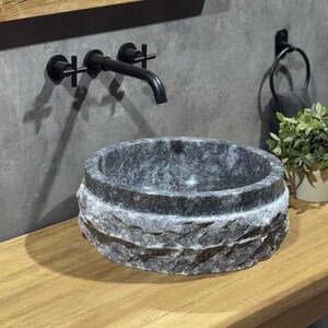 May include: A black faucet with a cross-shaped handle is mounted on a grey wall above a round, grey stone sink. The sink has a rough, textured exterior and a smooth, polished interior.