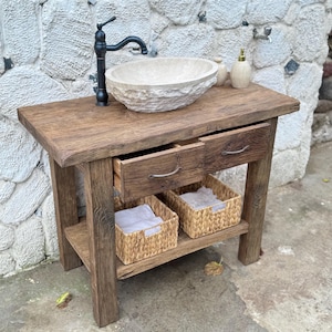 May include: A rustic wooden bathroom vanity with two drawers and a lower shelf. The vanity features a stone sink and a black faucet. Two woven baskets are on the shelf.