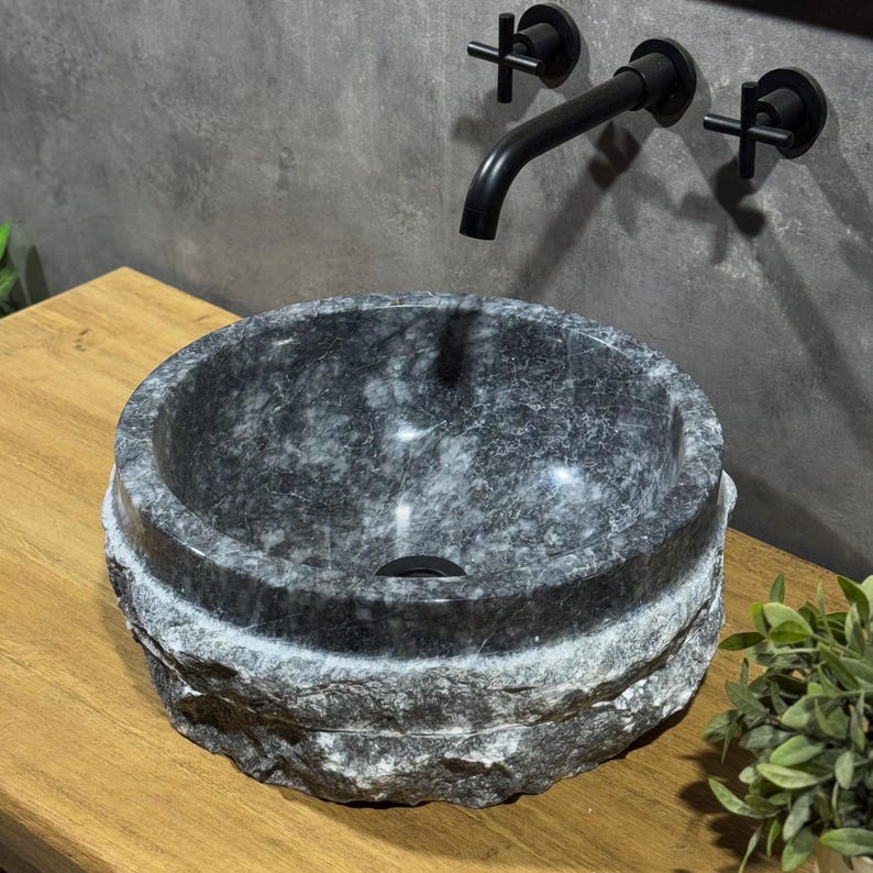 May include: A round, grey stone sink with a rough, textured exterior. The sink is set on a wooden countertop and has a black faucet with a modern design.