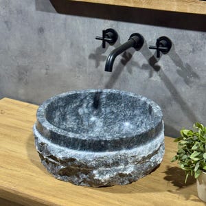 May include: A round, grey stone sink with a rough, textured exterior. The sink is mounted on a wooden countertop and has a black faucet with cross handles.