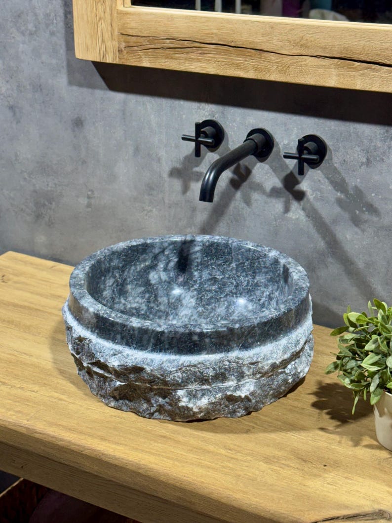 May include: A round, grey stone sink with a rough, textured exterior. The sink is mounted on a wooden countertop and has a black faucet with cross handles.
