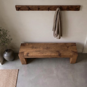 May include: A rectangular wooden bench with a natural grain finish and thick block legs. Above the bench, a long wooden wall-mounted coat rack features multiple pegs, with a single beige garment hanging from one of the hooks on the right side.