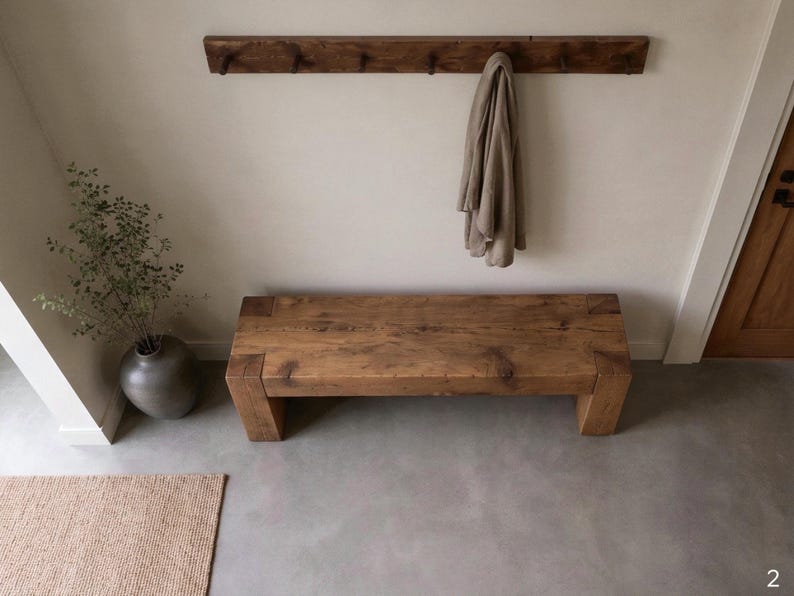May include: A rectangular wooden bench with a natural grain finish and thick block legs. Above the bench, a long wooden wall-mounted coat rack features multiple pegs, with a single beige garment hanging from one of the hooks on the right side.