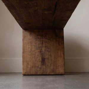 May include: A low-angle view of a heavy wooden bench featuring a thick, rectangular support leg and a wide, flat seat. The wood displays a deep brown tone with a prominent, natural grain pattern, visible knots, and a vertical crack running down the centre.