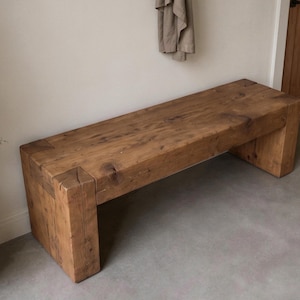 May include: A rectangular bench constructed from thick, solid brown wood with a visible grain and natural knots. The design features a flat seat supported by two sturdy, block-style legs at each end, showcasing a simple, heavy-duty construction.