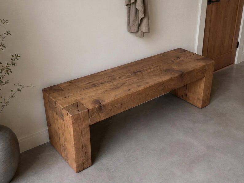 May include: A rectangular bench constructed from thick, solid brown wood with a visible grain and natural knots. The design features a flat seat supported by two sturdy, block-style legs at each end, showcasing a simple, heavy-duty construction.
