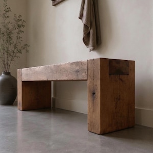 May include: A rectangular bench constructed from thick, reclaimed brown timber. The piece features a solid, block-style design with a flat seat and two sturdy, vertical support legs at each end, showcasing a natural wood grain and weathered texture.