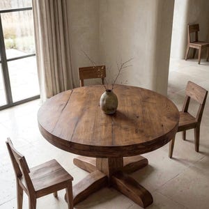 May include: A round wooden dining table with a thick top and a cross-shaped pedestal base, accompanied by three matching wooden chairs. A small, textured, earth-toned ceramic vase containing thin, bare branches sits in the center of the table.