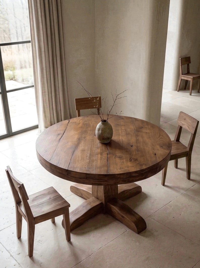 May include: A round wooden dining table with a thick top and a cross-shaped pedestal base, accompanied by three matching wooden chairs. A small, textured, earth-toned ceramic vase containing thin, bare branches sits in the center of the table.