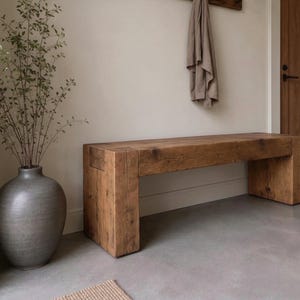 May include: A rectangular wooden bench with a natural grain finish and thick block legs, positioned next to a large, round, dark grey ceramic vase containing a tall, slender green plant with small leaves and thin, branching stems.