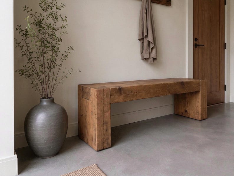 May include: A rectangular wooden bench with a natural grain finish and thick block legs, positioned next to a large, round, dark grey ceramic vase containing a tall, slender green plant with small leaves and thin, branching stems.