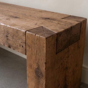 May include: A rectangular bench constructed from thick, reclaimed brown timber. The piece features visible wood grain, natural knots, and a sturdy joinery design where the horizontal seat meets the vertical leg with a flush, interlocking corner.