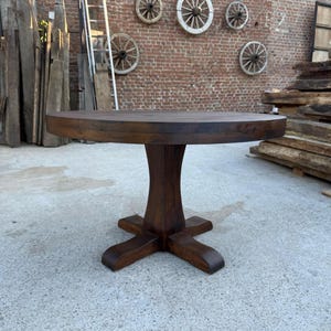 May include: A round dark brown wooden table with a thick tabletop and a central pedestal base featuring four curved feet. The table has a smooth, polished finish and a sturdy construction, standing on a flat, light-colored concrete surface.